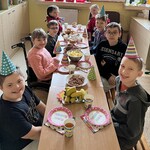 Uczniowie w kolorowych czapeczkach siedzą przy urodzinowym stole.