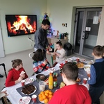 Opiekun częstuje uczennice opłatkiem. Wokół stołu wigilijnego siedzą uczniowie.