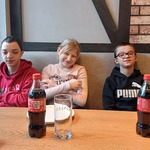 Trójka uczniów siedzi za stołem w pizzerni. Przed nimi na stole stoją talerze oraz butelki z colą.