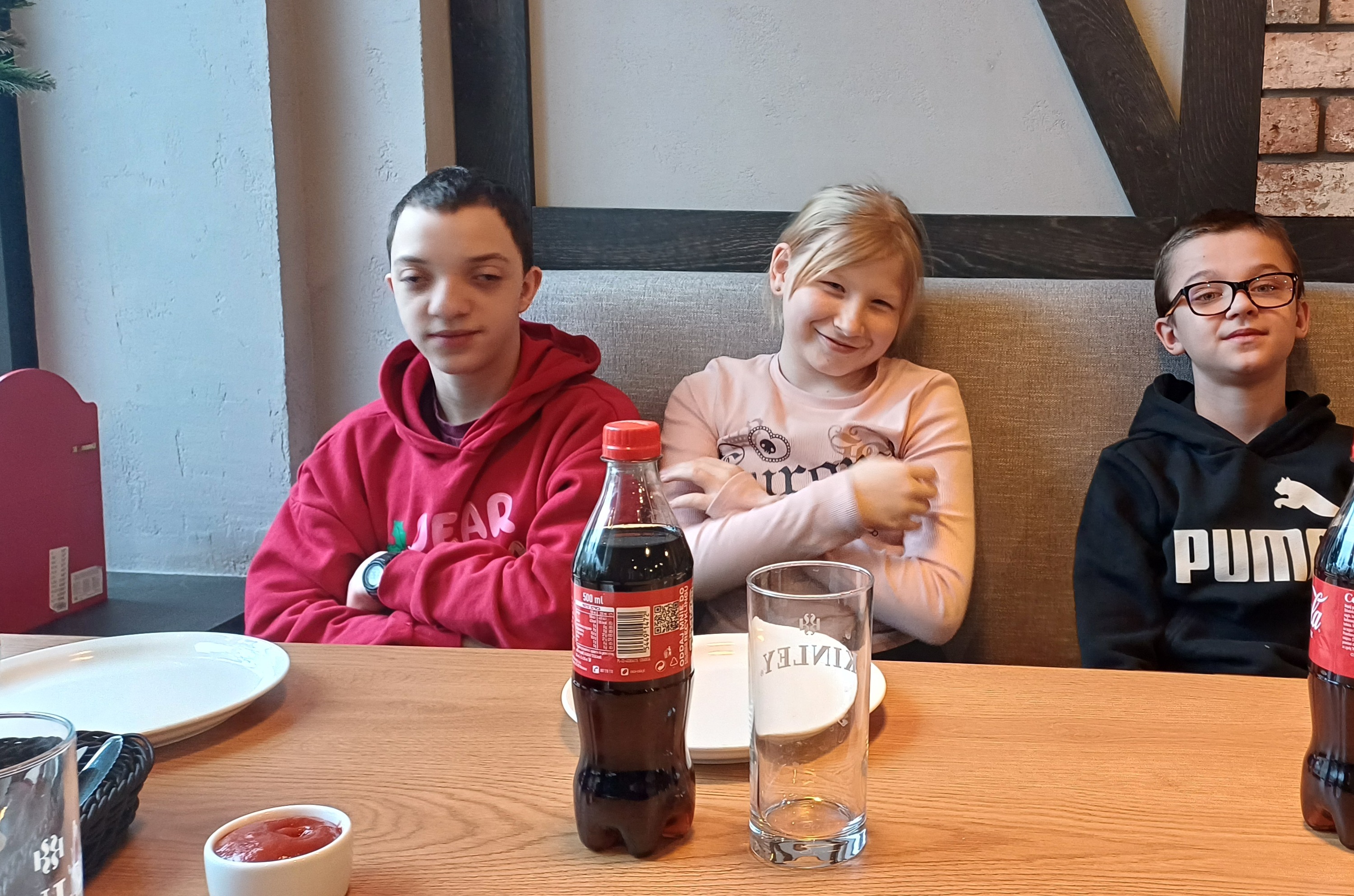 Trójka uczniów siedzi za stołem w pizzerni. Przed nimi na stole stoją talerze oraz butelki z colą.
