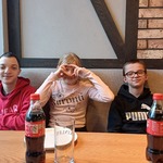 Trójka uczniów siedzi za stołem w pizzerni. Przed nimi na stole stoją talerze oraz butelki z colą.