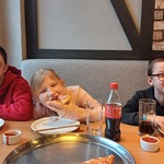 Trójka uczniów siedzi za stołem w pizzerni. Uczennica je pizzę.