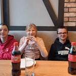 Trójka uczniów siedzi za stołem w pizzerni. Przed nimi na stole stoją talerze oraz butelki z colą.