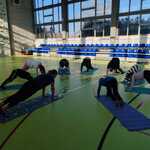 Na sala sportowej nauczycielka i uczniowie wykonują ćwiczenia gimnastyczne na matach.