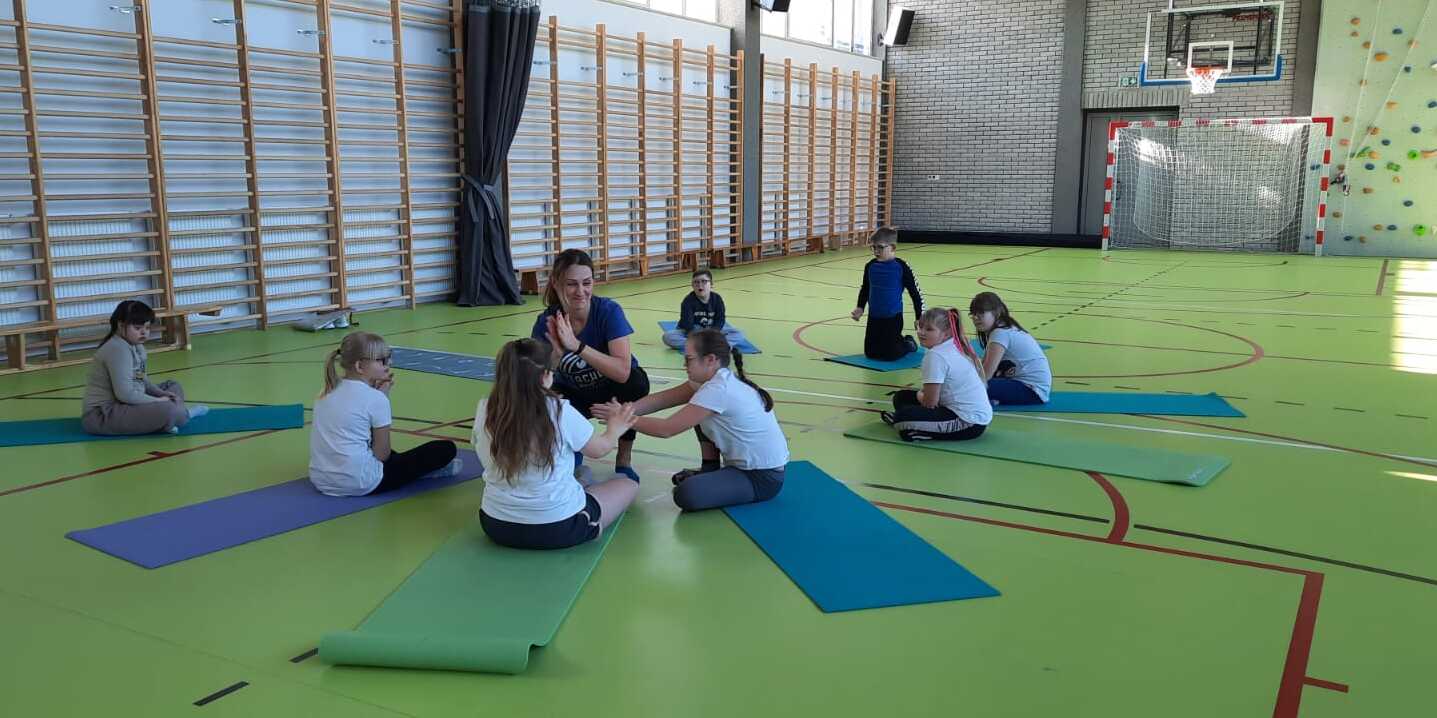 Na sala sportowej nauczycielka i uczniowie wykonują ćwiczenia gimnastyczne na matach.