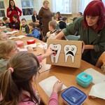 Uczenica wkłada karteczkę w otwór pudełka z rysunkiem zębów. Pudeko trzyma nauczycielka.