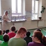 Nauczycielka mówi do uczniów siedzących na ławeczkach.