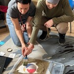 Nauczycielka trzyma ucznia za rękę pomagając mu pędzlem odgarnąć kamyczki z obrazka