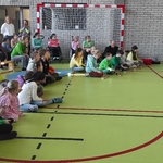 Uczniowie siedzą na ławeczkach i podłodze. Uczniowie siedzący na podłodze grają na cymbałkach. Nauczycielka pokazuje kolorowe karteczki na patykach.