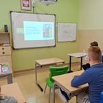 Uczniowie siedzą przy stolikach i oglądają slajdy wyświetlane na ekranie.