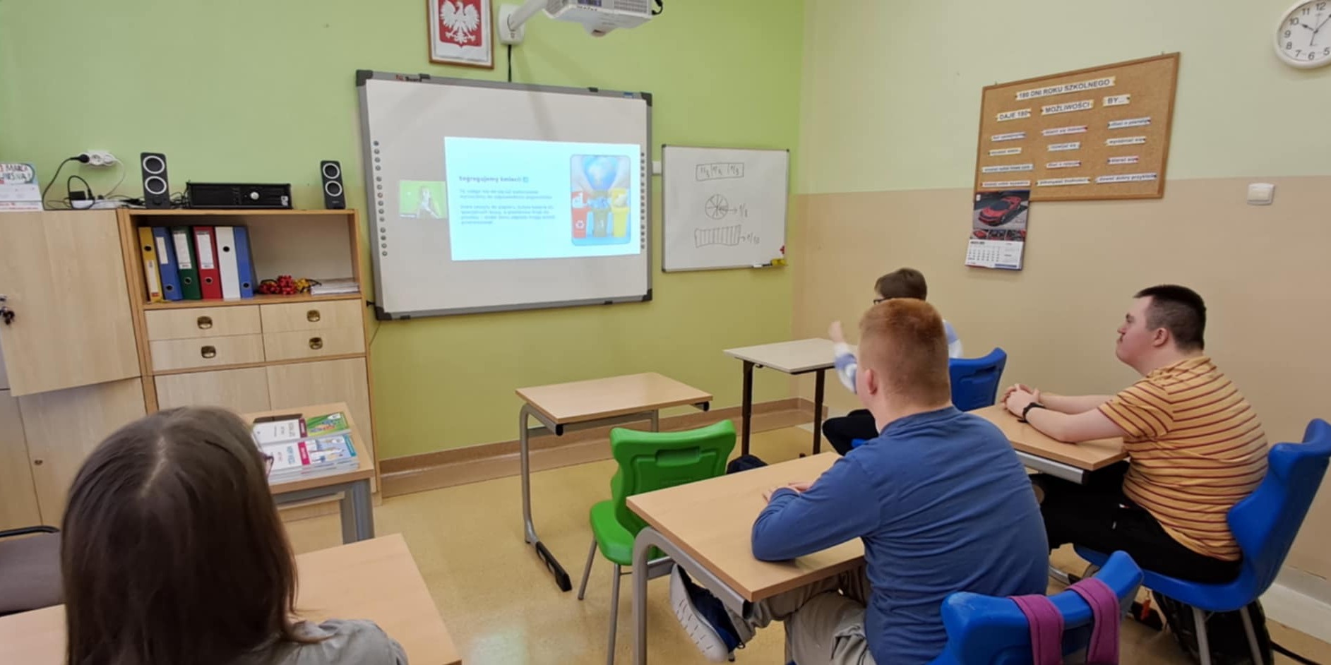 Uczniowie siedzą przy stolikach i oglądają slajdy wyświetlane na ekranie.