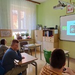Uczniowie siedzący przy stolikach i  stojący pod oknem oglądają slajdy wyświetlane na ekranie.