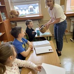 Nauczycielka rozdaje słodkości z koszyczka uczniom siedzącym w ławkach.