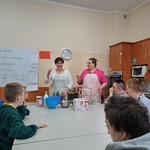 Uczniowie siedzący wokół stołu obserwują nauczycielki, przed którymi leżą na stole produkty spożywcze.