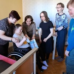 Natalka siedzi na wózku. Obok stoi jej mama. Trójka uczniów obserwuje nauczycielkę , która podaje dziewczynce na wózku prezent.