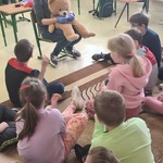 Nauczycielka z misiem na kolanach mówi do dzieci siedzących przed nią na podłodze.