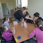 Grupa uczniów siedzi wokół stołu. Nauczycielka podaje zapach do powąchania uczniowi.