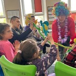 Nauczyciel w kolorowej peruce podaje uczennicy kolorowy kwiat