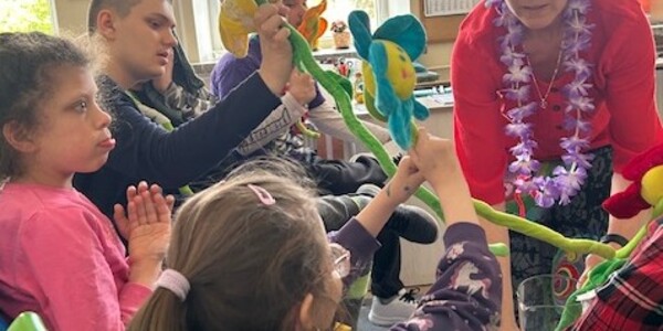 Nauczyciel w kolorowej peruce podaje uczennicy kolorowy kwiat
