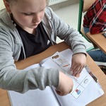 Uczennica siedzi przy stoliku. Wkleja kartę pracy do zeszytu.