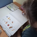 Uczennica siedzi przy stoliku. Wkleja obrazki do zeszytu.