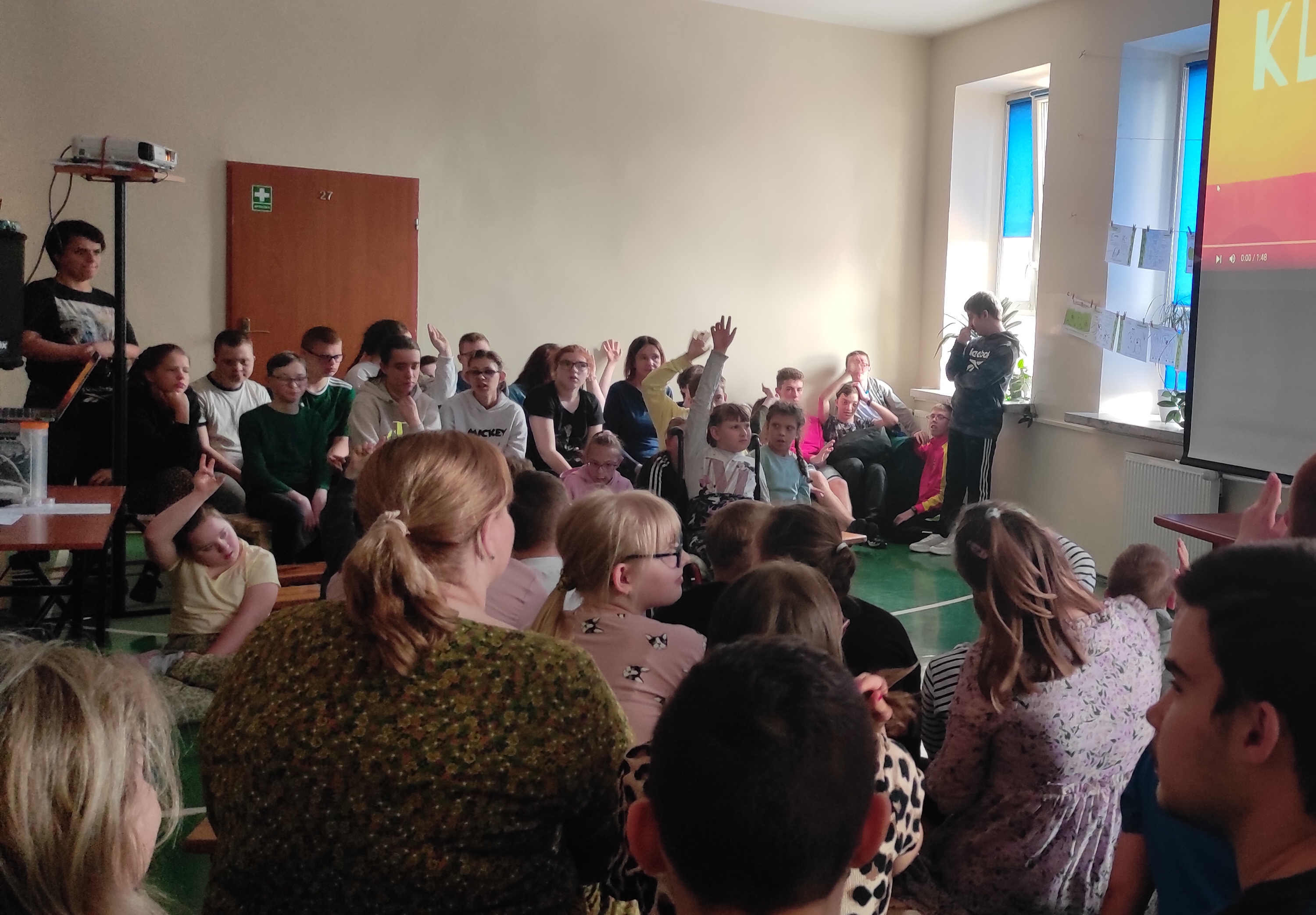 Nauczycielka omawia slajd wyświetlany na ekranie. Na ławkach siedzą uczniowie