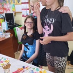 Jubilatka siedzi za stołem obok stoi nauczycielka i uczennica