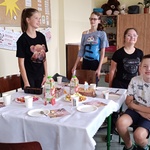 Jubilat siedzi przy urodzinowym stole wokół którego stoją koleżanki