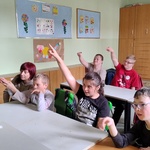Uczniowie siedzą w ławkach  z podniesioną ręką do odpowiedzi