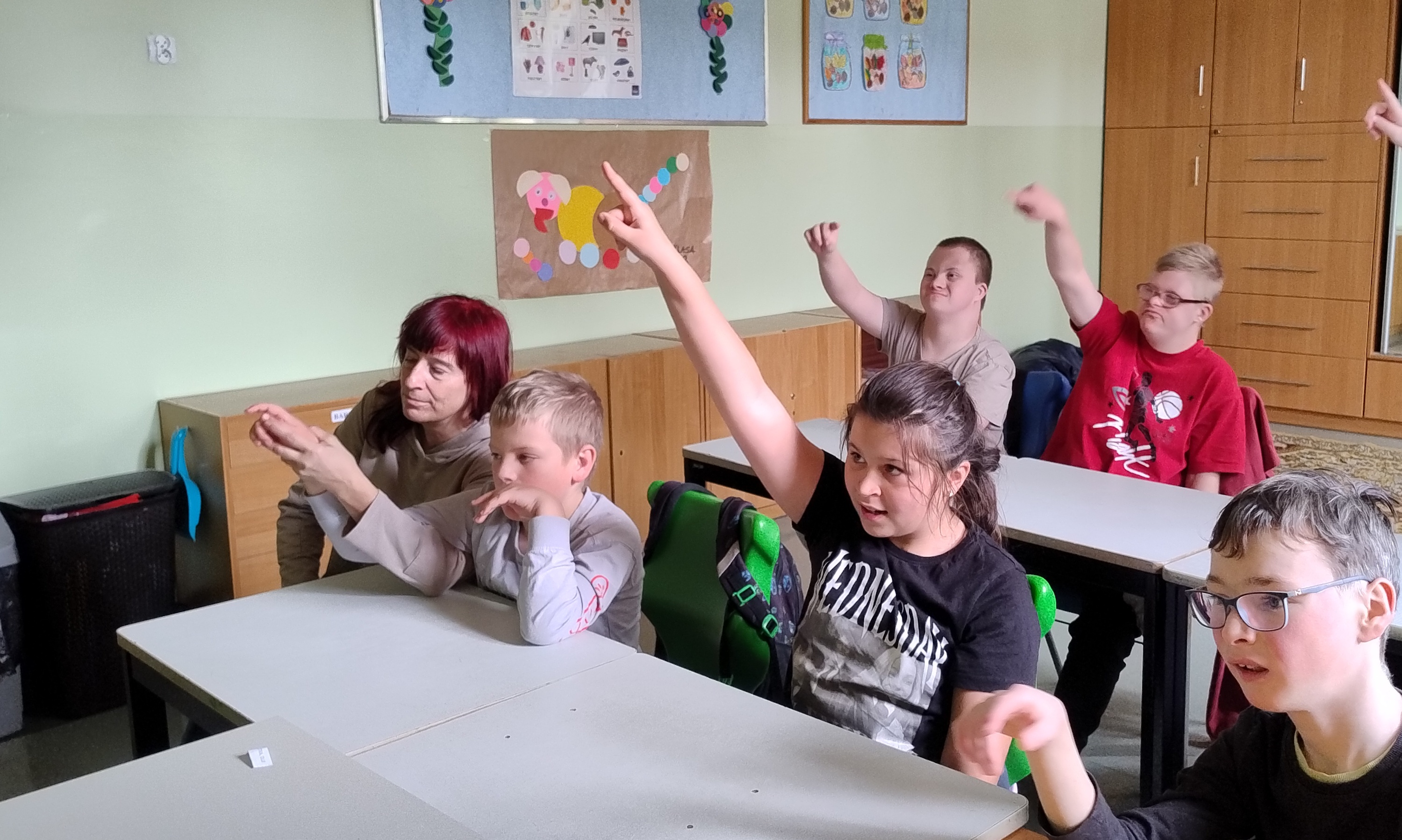 Uczniowie siedzą w ławkach  z podniesioną ręką do odpowiedzi
