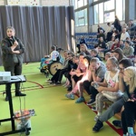 Pani edukatorka mówi przez mikrofon stojąc przed uczniami