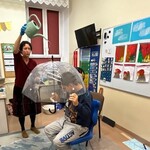 Uczeń siedzi na krześle pod parasolem, nauczyciel leje wodę konewką