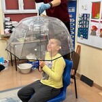 Uczeń siedzi na krześle pod parasolem, nauczyciel leje wodę konewką
