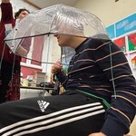 Uczeń siedzi na krześle pod parasolem, nauczyciel leje wodę konewką
