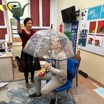 Uczeń siedzi na krześle pod parasolem, nauczyciel leje wodę konewką