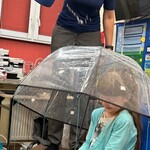 Uczennica siedzi na krześle pod parasolem, nauczycielka leje wodę konewką