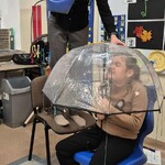 Uczennica siedzi na krześle pod parasolem, nauczycielka leje wodę konewką