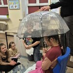 Uczennica siedzi na krześle pod parasolem, nauczycielka leje wodę konewką