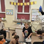 Uczeń siedzi na krześle pod parasolem, nauczycielka leje wodę konewką, uczniowie podczas zajęć