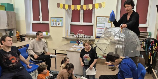 Uczeń siedzi na krześle pod parasolem, nauczycielka leje wodę konewką, uczniowie podczas zajęć