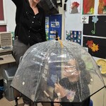 Uczennica siedzi na krześle pod parasolem, nauczycielka leje wodę konewką