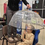 Uczennica siedzi na krześle pod parasolem, nauczycielka leje wodę konewką