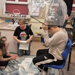 Uczeń siedzi na krześle pod parasolem, nauczycielka leje wodę konewką