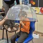 Uczeń siedzi na krześle pod parasolem, nauczycielka leje wodę konewką