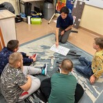 Nauczyciel pokazuje palcem na plan zajęć, uczniowie siedzą na podłodze, obserwują.