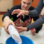 Uczeń wsypuje mąkę do miski. Nauczyciel pomaga przytrzymując torbę z mąką