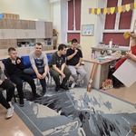 Uczniowie siedzą na krzesłach. Przednimi stoi nauczycielka z kartkami w rękach