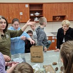 Pani edukatorka pokazuje uczniom stojącym wokół stołu wykonaną pyzę dla ptaków