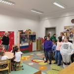 Grupa uczniów stoi w sali przedszkolnej. Przed nimi przy stolikach siedzą przedszkolaki