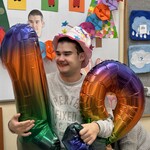 Uczeń siedząc na krześle trzyma duże nadmuchane balony-cyfry: 1 i 9